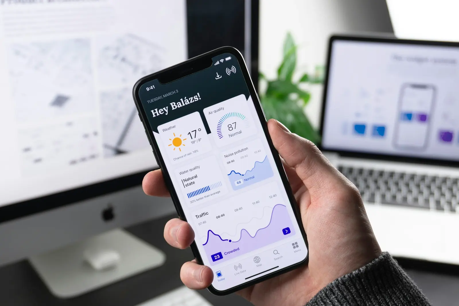 Hand holding a phone with a mobile app open, a laptop with app screen designs behind it